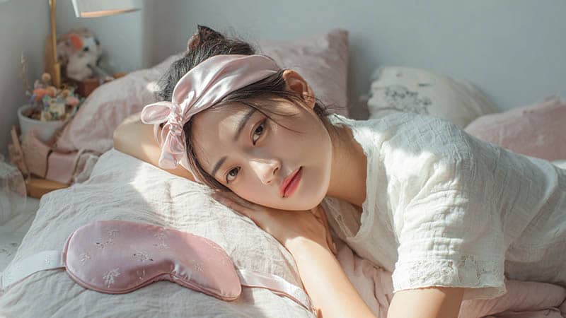 A woman using a silk heatless hair curler and silk headband, with a mulberry silk eye mask on the bed.