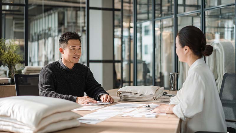 Buyer negotiating silk manufacturing prices with a factory, with silk pillowcase and silk sleep mask samples on the table, showing quality control documents and cost discussion.